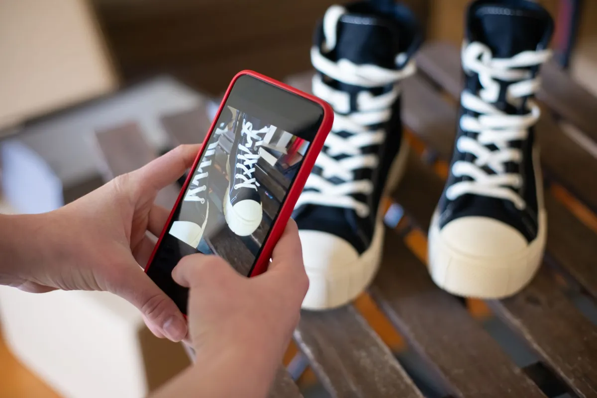 Hands using a smartphone to photograph black sneakers on a wooden surface