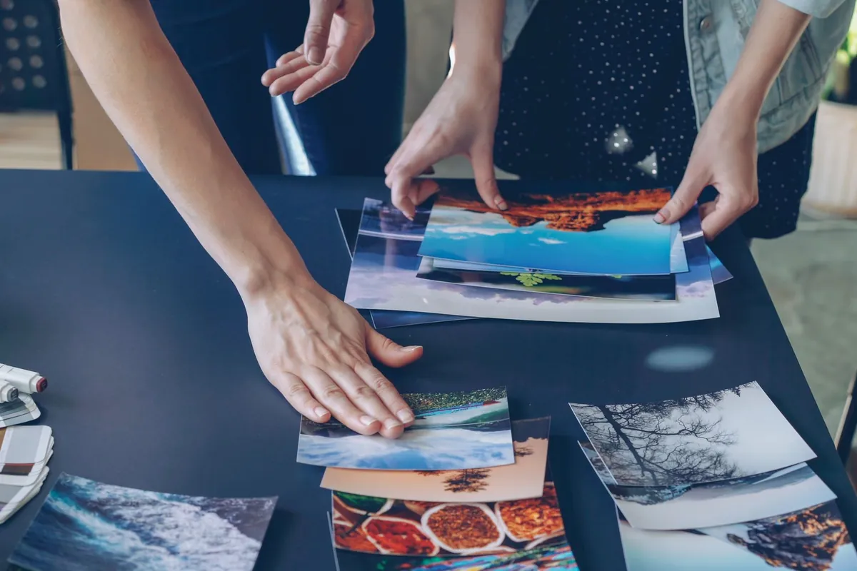 Two people's hands sorting printed photos on a dark table, like a brand team choosing which creator videos to run as Spark Ads